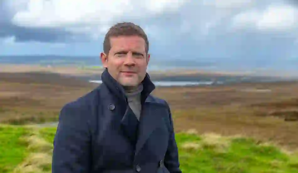 Dermot O'Leary standing in windswept landscape at Malin Head, with rolling hills, water and dramatic clouds in the background.