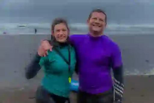 Dermot O'Leary and surfer in wetsuits smiling on Strandhill Beach after surfing, with waves and misty shoreline behind.