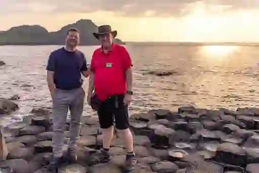 Dermot O'Leary and a storyteller standing on basalt columns at the Giant’s Causeway at sunset, with sea views and rugged coastline behind.