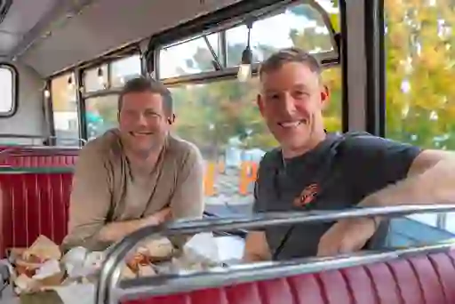 Dermot O'Leary and owner seated inside Pyke ’n’ Pommes in Derry~Londonderry, enjoying loaded chips at a retro bus-style dining booth.