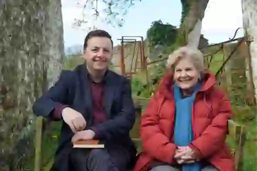 Sandi Toksvig and Oliver Callan smile on a wooden bench between old stone walls, with a traditional farm gate behind them.
