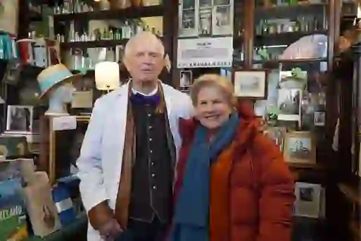 Sandi Toksvig smiles beside a man in a white coat inside historic Sweny's Pharmacy filled with books and memorabilia.