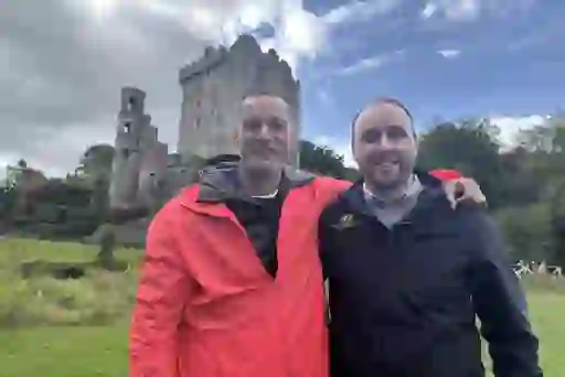 Fred and a tour guide smiling in front of Blarney Castle, with the medieval stone tower rising above landscaped gardens in County Cork.
