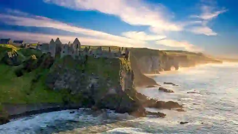 Ruins of Dunluce Castle, County Antrim, on a steep cliff above the Atlantic, with rugged coastline, sea arches and soft golden light at sunset.
