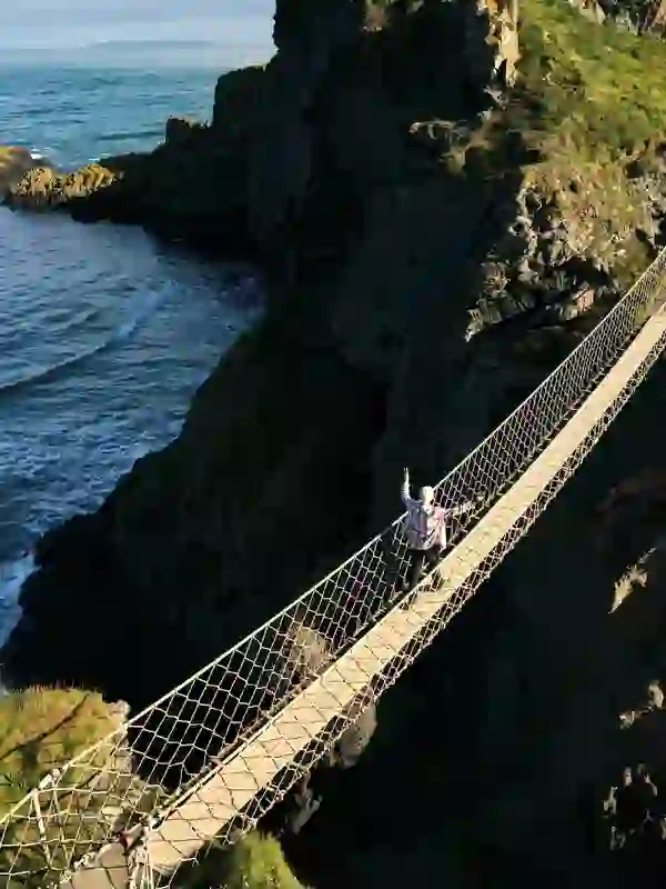 Person crossing the Carrick-a-Rede rope bridge above a rocky coastline with waves breaking below in County Antrim.