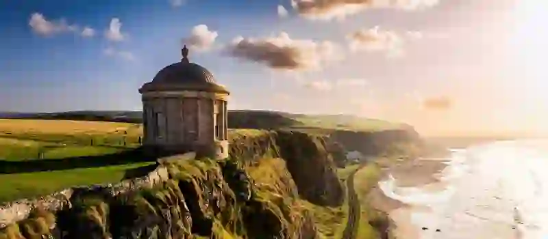 Mussenden Temple stands on a cliff in County Londonderry, overlooking Downhill Beach and golden fields at sunset.
