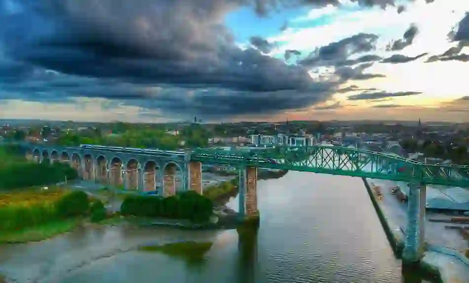 A train crosses the Boyne Viaduct at sunset, with dramatic clouds over the River Boyne and the town of Drogheda in the background.