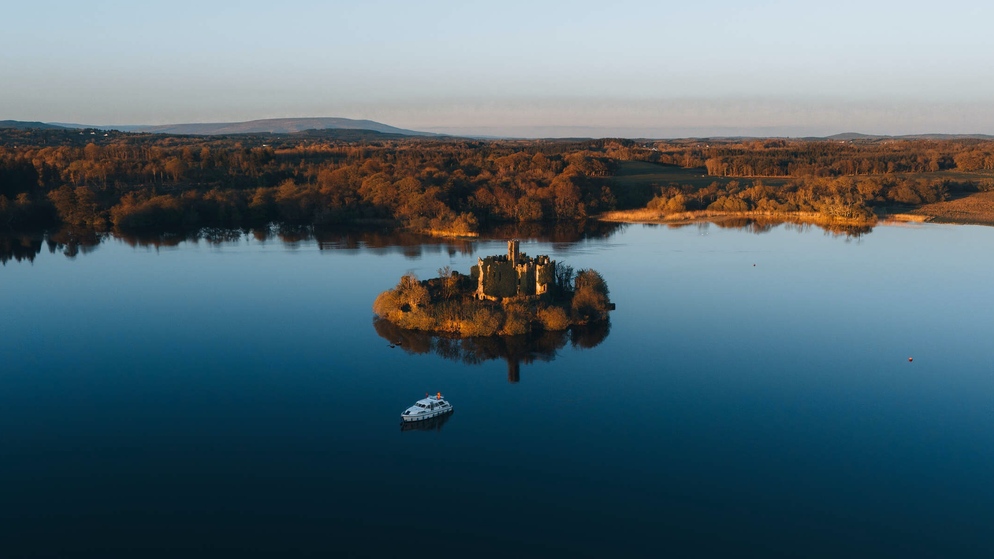 Autumn trees line the banks of a quiet lake, with a wooded island and a boat below.