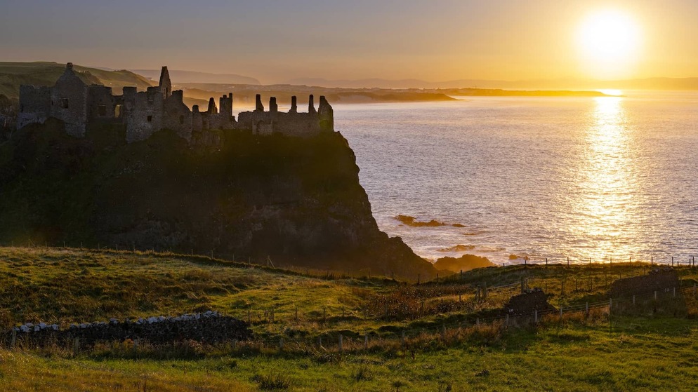 Sunset over Dunluce Castle ruins on the Causeway Coast, with golden light reflecting across the Atlantic Ocean.