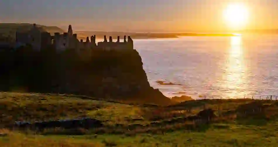 Sunset over Dunluce Castle ruins on the Causeway Coast, with golden light reflecting across the Atlantic Ocean.