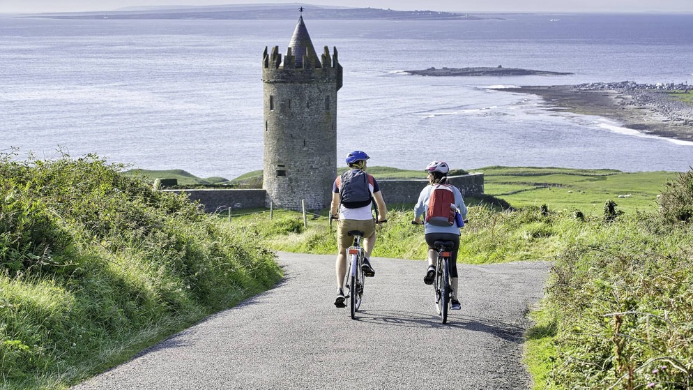 Two cyclists approach Doonagore Castle near Doolin, County Clare, with views of the Atlantic Ocean beyond.