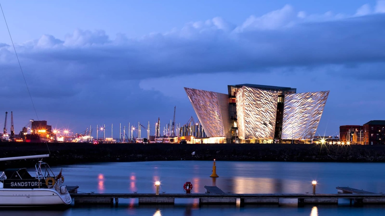 Titanic Belfast illuminated at dusk beside the River Lagan, with marina boats and city lights reflecting on the water.