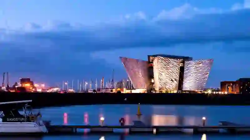 Titanic Belfast illuminated at dusk beside the River Lagan, with marina boats and city lights reflecting on the water.