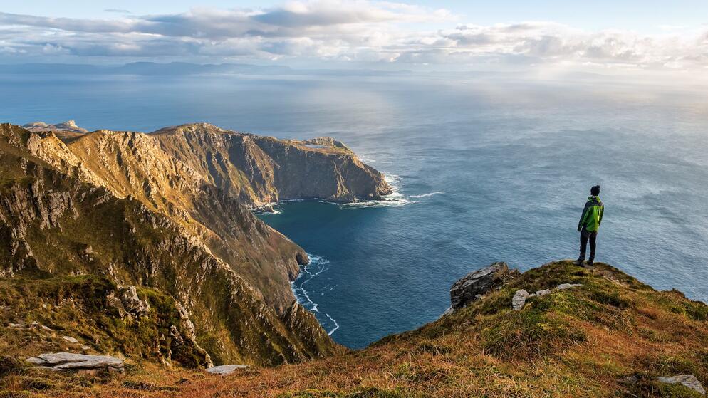 Una persona con una giacca verde sta in piedi sul bordo di una scogliera che si affaccia sulle scogliere di Slieve League e sull'Oceano Atlantico nella contea di Donegal.