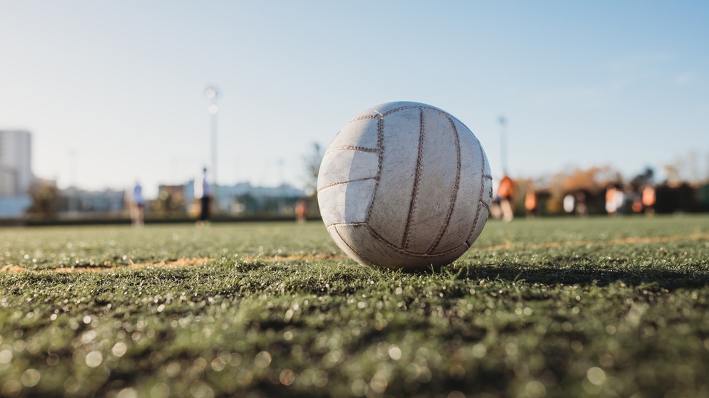 Fútbol gaélico en un campo iluminado por el sol con jugadores desdibujados entrenando al fondo en un campo de la GAA.