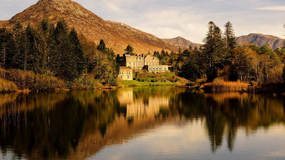 Ballynahinch Castle nestled among trees by a still lake, with rugged mountains rising in the background.