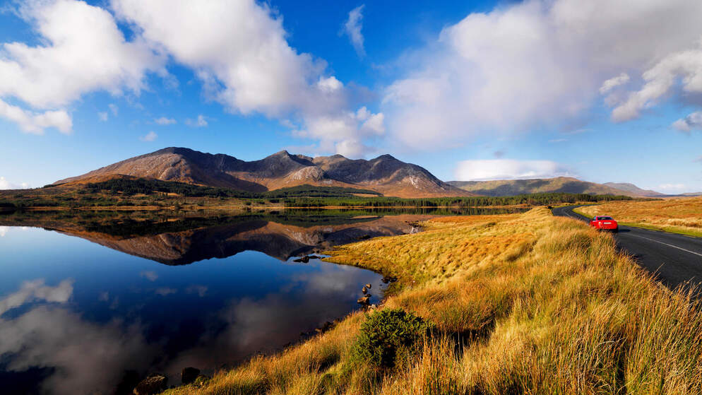 Rotes Auto, das entlang einer ruhigen Straße an einem spiegelglatten See und einer Bergkette in Connemara, Grafschaft Galway, fährt.