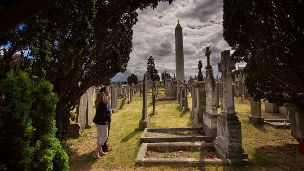 Visitors walking among historic gravestones at Glasnevin Cemetery, Dublin, with the tall round tower in the background.