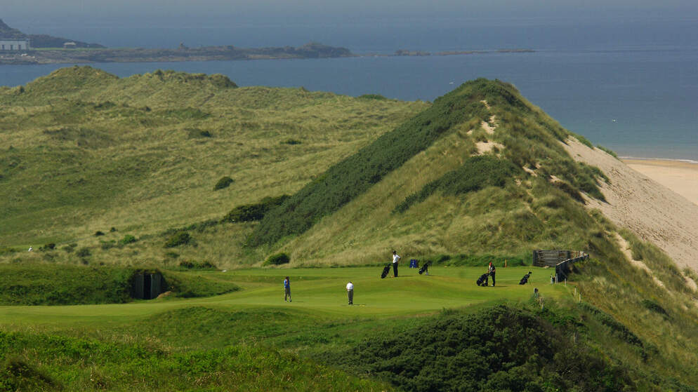 Sand dunes and fairways meet the Atlantic coast in a wide view of an Irish links course under blue skies.