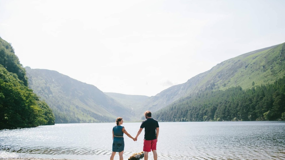 Couple holding hands by a lake at Glendalough, County Wicklow, framed by forested mountains under a pale sky.