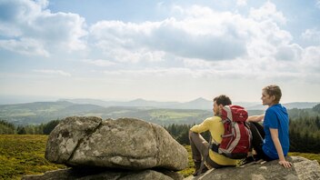 Two hikers with backpacks enjoy scenic views from a rocky hilltop in rural Ireland under a cloudy sky.