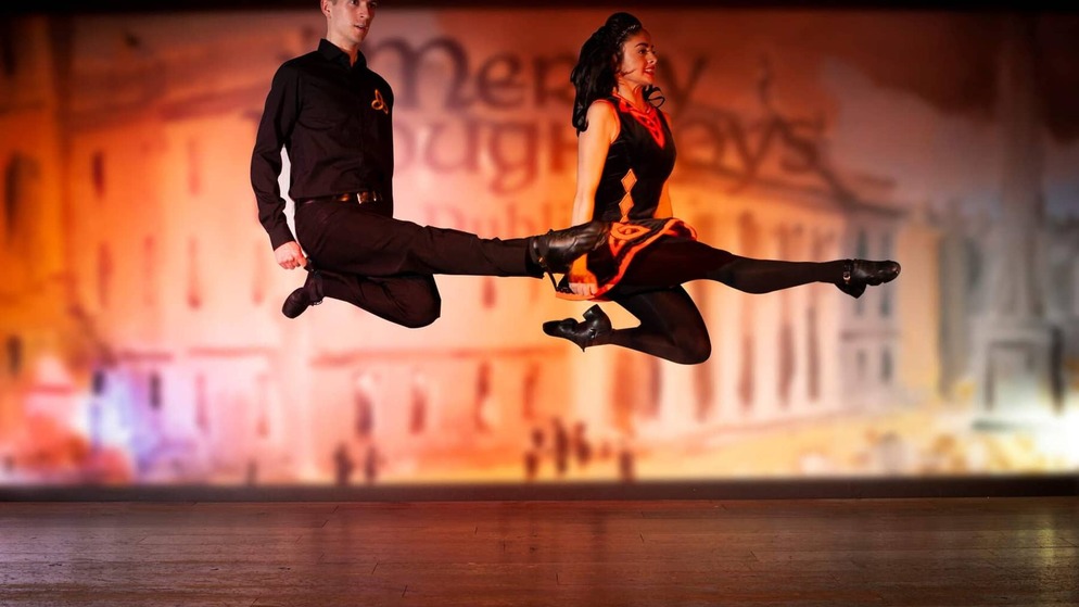 Bailarines irlandeses con trajes negros y rojos danzan en el aire durante una actuación de baile tradicional.