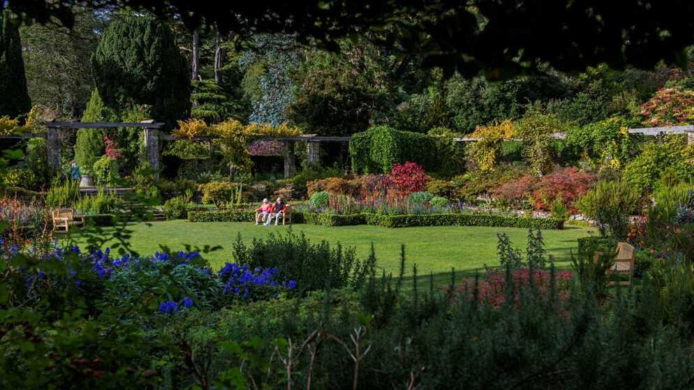 Sunlit gardens at Mount Stewart with colourful flowerbeds, manicured lawns, and visitors relaxing on benches.