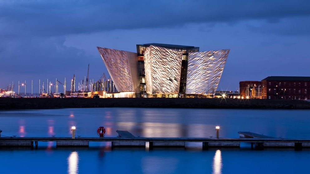 Titanic Belfast lit up at dusk, its angular facade reflected in the water, with a dark blue sky behind.