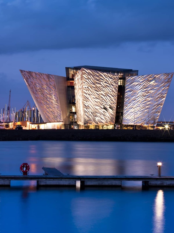 Titanic Belfast illuminato al tramonto, la sua facciata angolare riflessa sull'acqua, con un cielo blu scuro sullo sfondo.