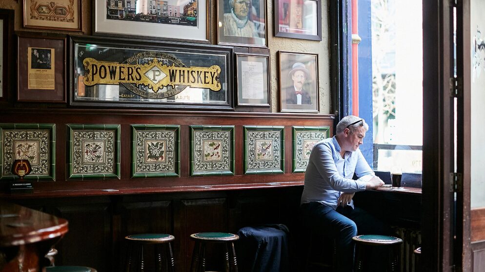 Un hombre lee junto a una ventana en un pub histórico de Dublín con retratos enmarcados y paneles de azulejos antiguos.