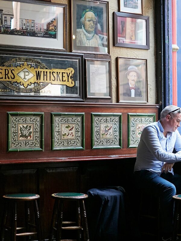 Man is aan het lezen naast een raam in een historische pub in Dublin met ingelijste portretten en vintage tegelpanelen.