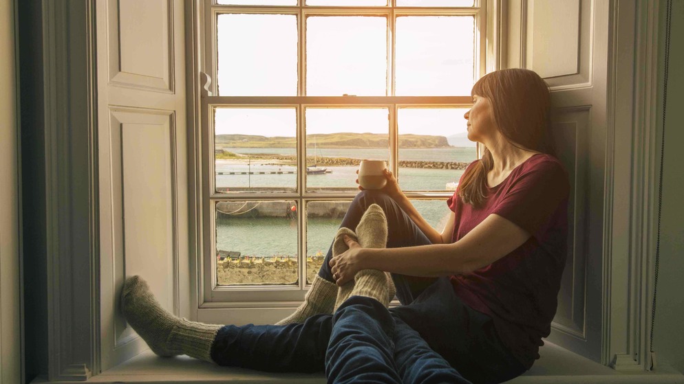 Eine Frau sitzt mit einem heißen Getränk an einem Sprossenfenster und beobachtet einen goldenen Sonnenuntergang über einem ruhigen Hafen auf der Insel Rathlin.