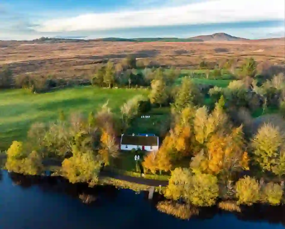 Keenaghan Cottage beside a lakeshore, surrounded by autumn trees, with rolling bogland and a distant mountain in County Fermanagh.