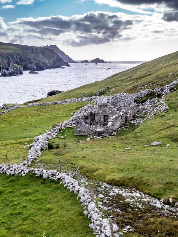 Ruined stone cottage on grassy slope above cliffs and sea on the Irish coast, under a cloudy sky