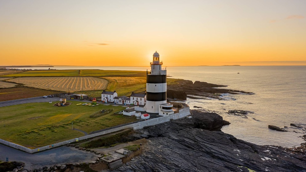 Hook Head Lighthouse glowing at sunset on the Wexford coast, beside cottages and golden fields by the Irish Sea.
