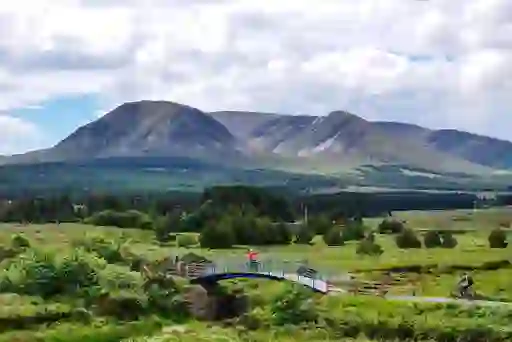 Fietsers die een brug oversteken op de Great Western Greenway in County Mayo, met groene velden en bergen op de achtergrond.