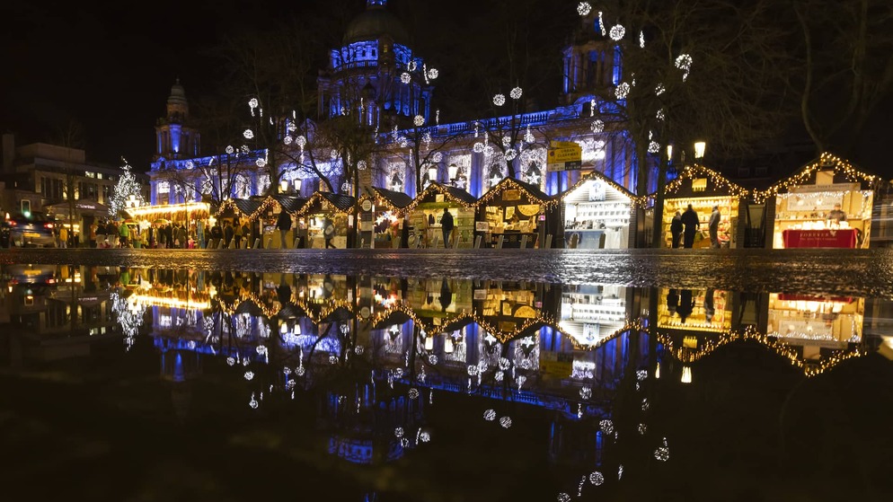 Christmas market huts glowing with fairy lights reflected in puddles before Belfast City Hall at night.