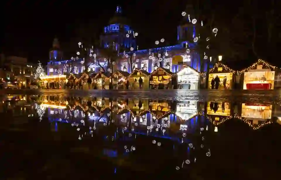 Christmas market huts glowing with fairy lights reflected in puddles before Belfast City Hall at night.