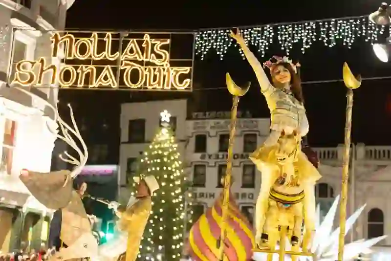 Corkmas-Festumzug in Cork mit Künstlern auf goldenen Festwagen unter einem „Nollaig Shona Duit”-Schild und einem beleuchteten Weihnachtsbaum im Hintergrund.