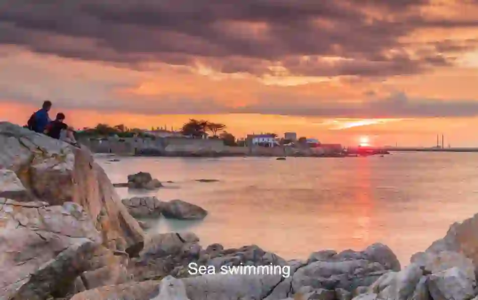 People watching the sunset over Dalkey, County Dublin, with rocky shoreline.