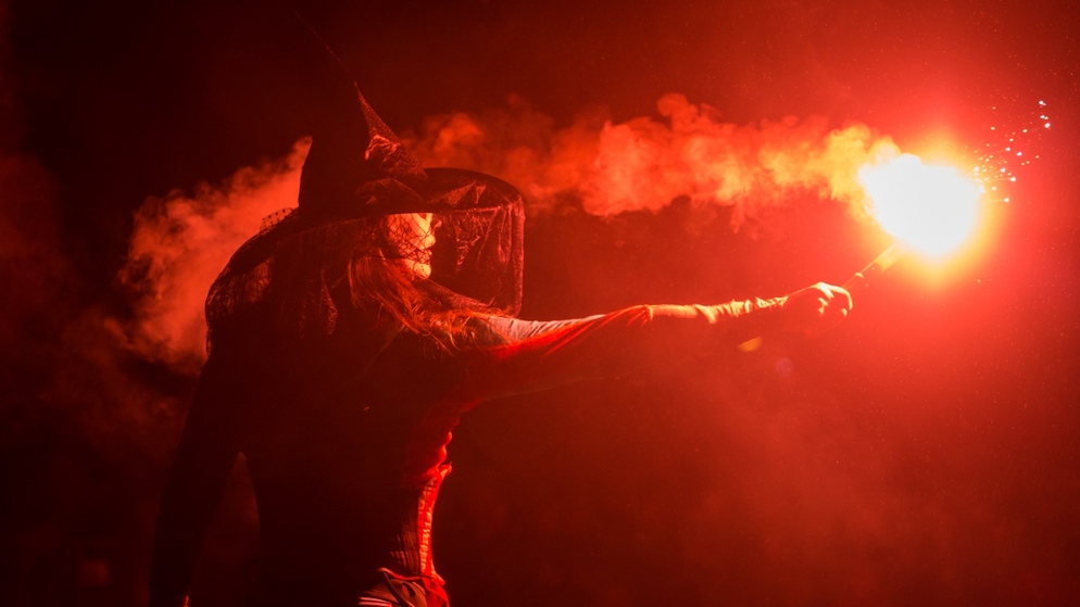 Frau in einem Hexenhut hält ein leuchtend rotes Leuchtfeuer in der rauchigen Dunkelheit während einer Halloween-Veranstaltung in Irland.