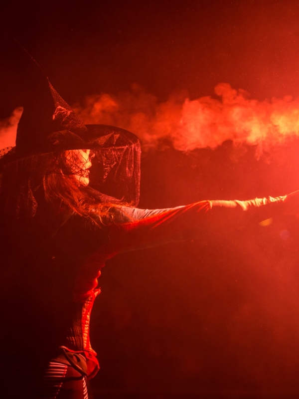 Woman in a witch’s hat holds a bright red flare in smoky darkness during a Halloween event in Ireland.