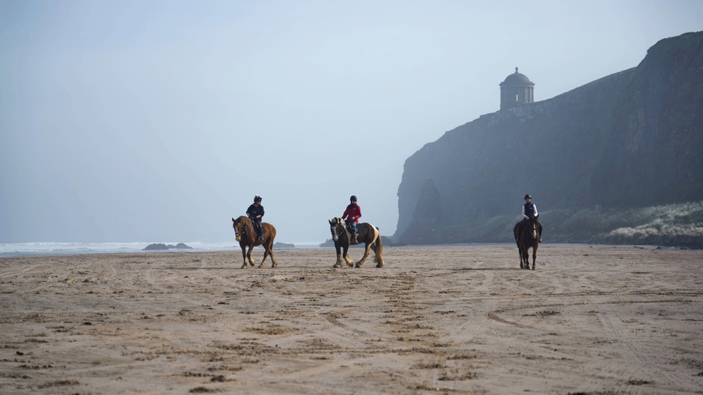 Three horse riders cross Downhill Beach, County Londonderry, with Mussenden Temple perched on the cliffs above in the mist.