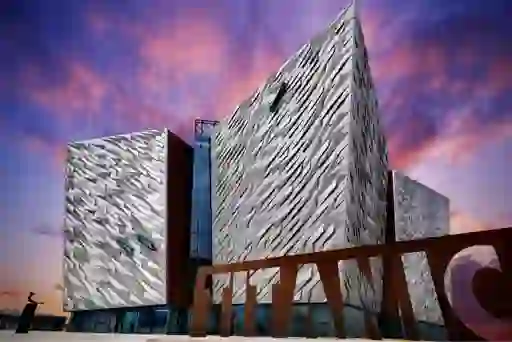 Titanic Belfast's angular, silver-clad exterior beneath a pink and purple sunset sky, with large TITANIC lettering in front.