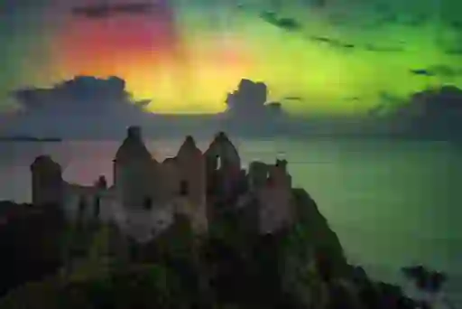 The ruins of Dunluce Castle in County Antrim perched on a rocky cliff above the sea, beneath colourful Northern Lights.