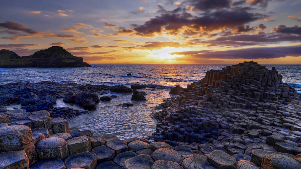 Hexagonal basalt columns at the Giant's Causeway in County Antrim, with waves washing against the rocks beneath a golden sunset.