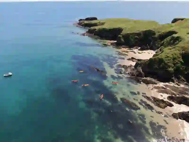 Kayakers paddle along a clear turquoise shoreline with rocky inlets and sandy coves on a rugged coast.