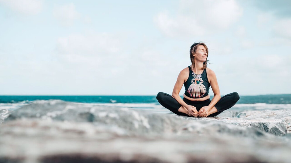 Eine Frau, die im Schneidersitz Yoga auf Felsen an der Küste von Irland macht.