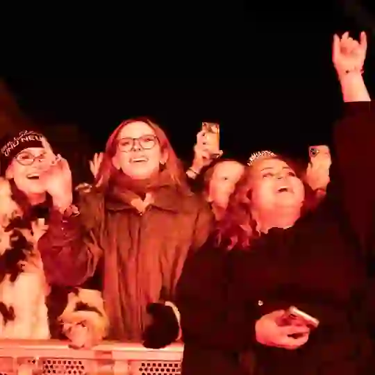 Festivalgoers cheering, smiling and raising their hands behind a crowd barrier at New Year's Festival Dublin, illuminated by bright red stage lighting.