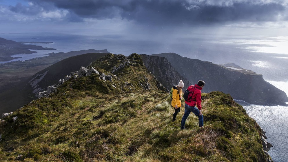 Two hikers in bright jackets walk along a cliff edge on Slieve League, overlooking the Atlantic Ocean.